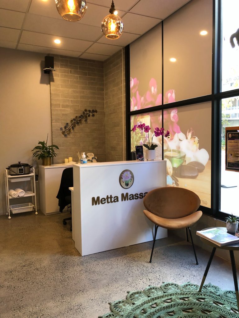 A bright and welcoming massage therapy reception area with a white counter, a comfortable waiting chair, and a large window with a floral design